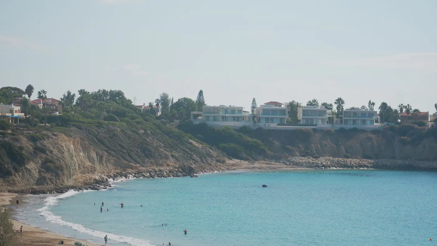 Beautiful beach scene with clear turquoise waters, sandy shore, and cliffside homes under a bright sky.
