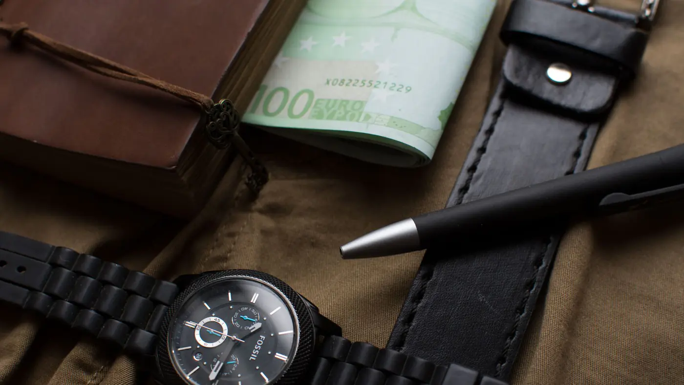 Flat lay featuring a watch, euros, leather belt, and a pen on a dark background.