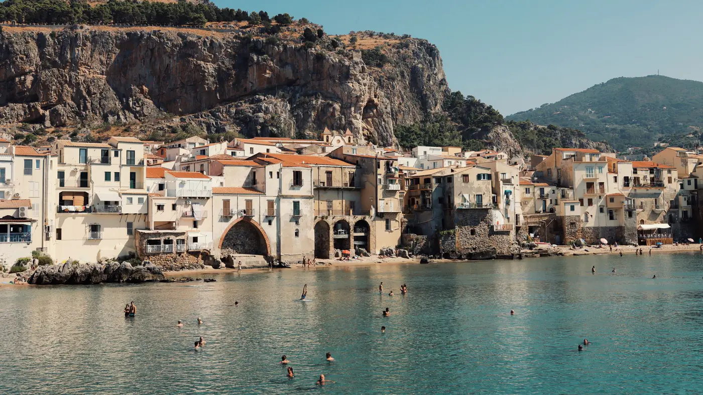 Swimmers enjoy the water with Cefalù's historic seaside buildings and rocky cliffs in Sicily, Italy.