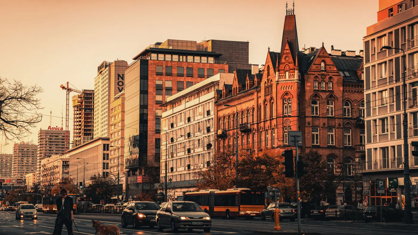 City street in Warsaw during sunset with historic buildings, traffic, and pedestrians.