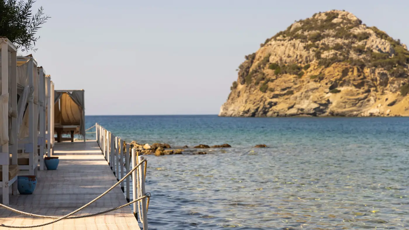 Relaxing view of a wooden pier extending into crystal-clear sea beside a rocky island during a sunny day.