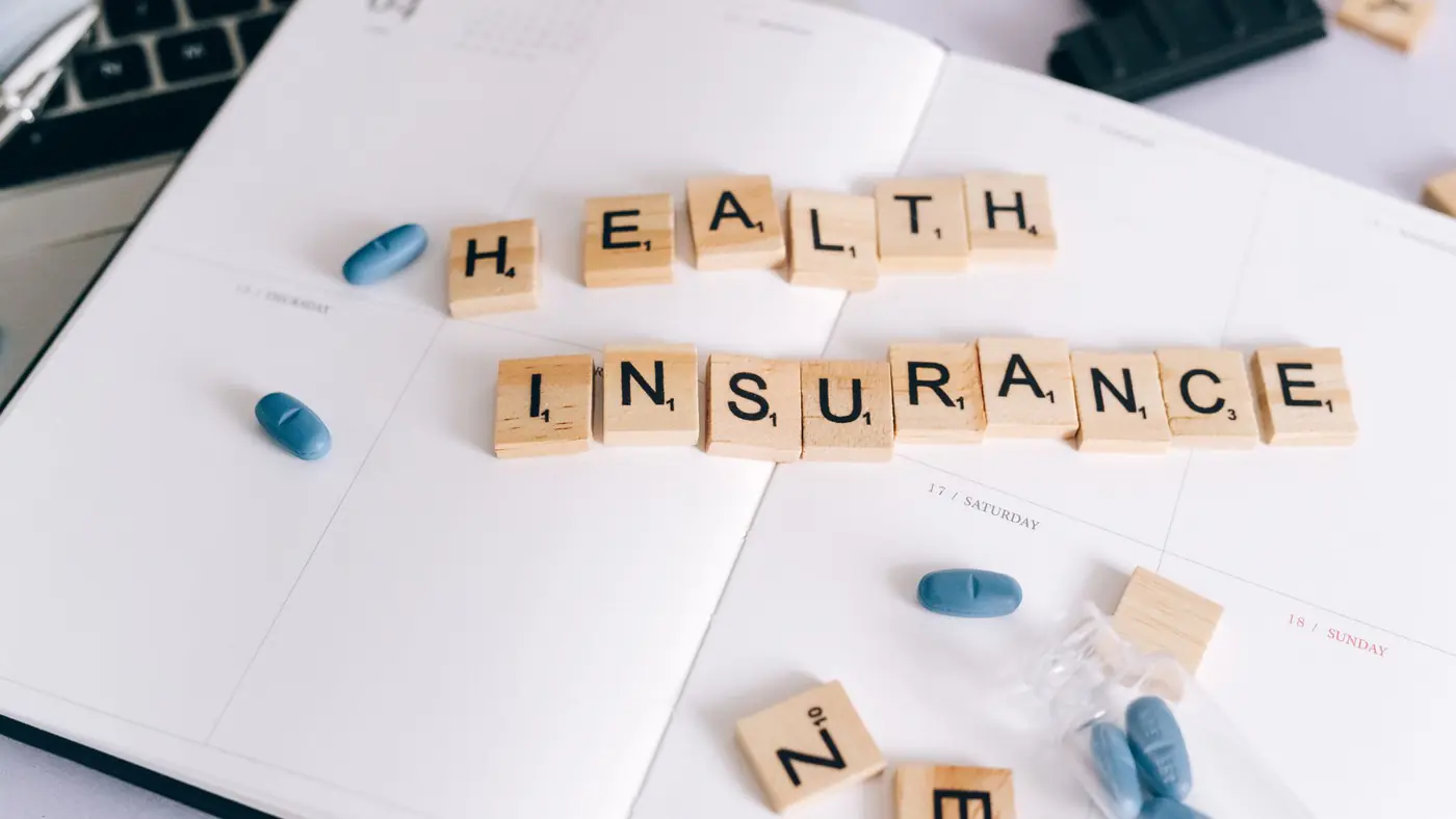 Scrabble tiles spelling 'Health Insurance' on planner with pills and laptop, symbolizing healthcare planning.