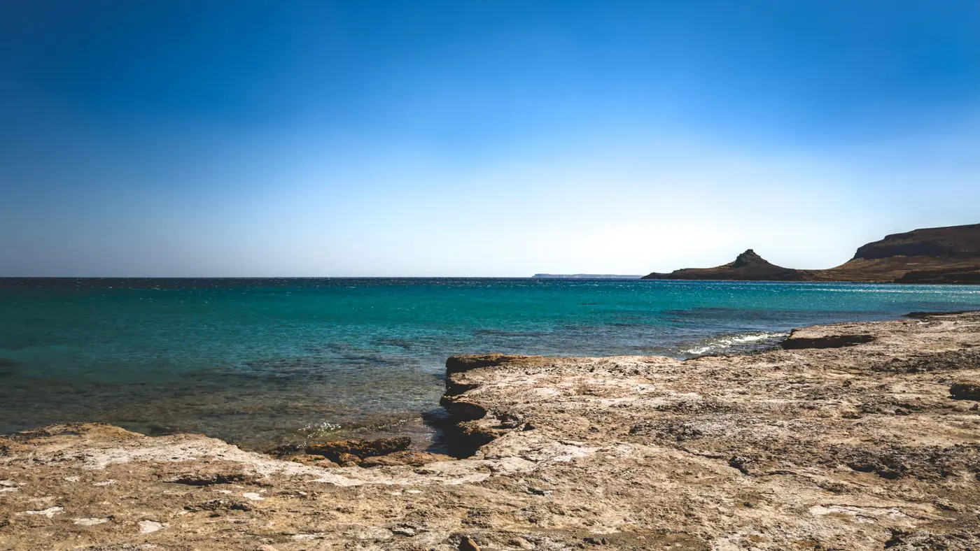A tranquil beach scene featuring a rocky shore, turquoise sea, and a clear blue sky.