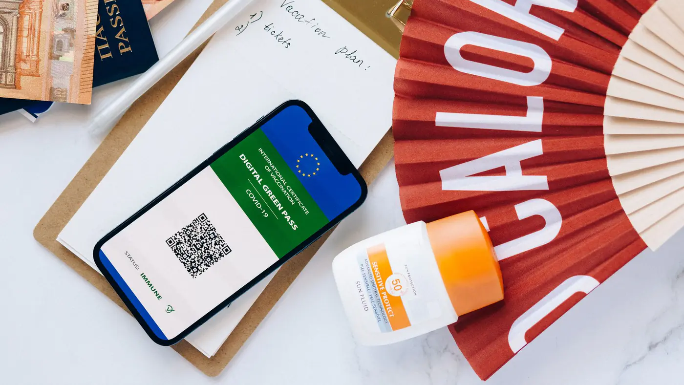 Flat lay of travel essentials featuring a digital green pass, passport, and other travel items on a table.