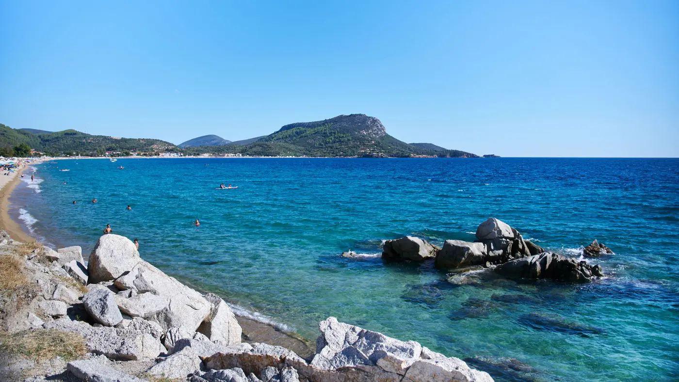 Idyllic beach scene in Greece with turquoise waters and rocky shoreline under clear blue sky.