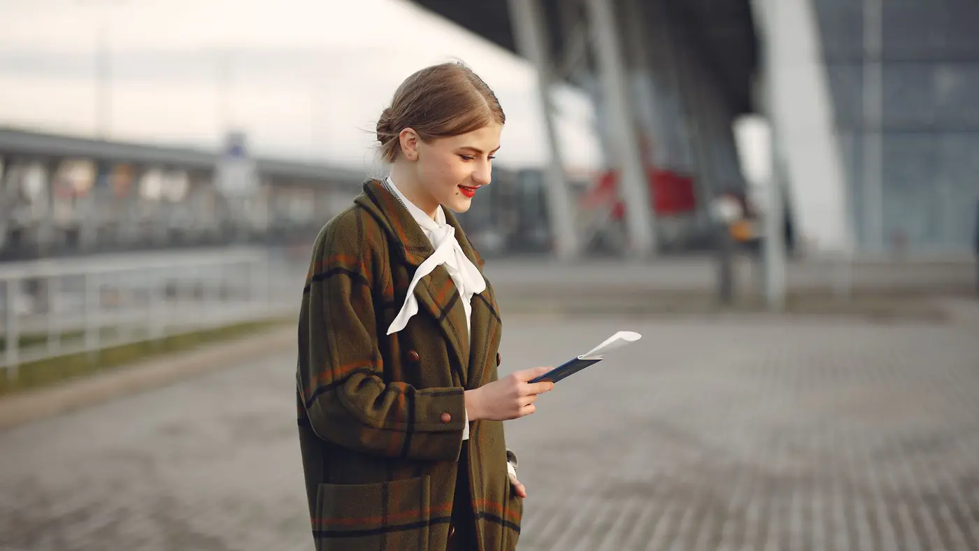 Smiling female passenger wearing trendy plaid coat and white blouse checking passport and ticket standing on pavement near contemporary building of airport
